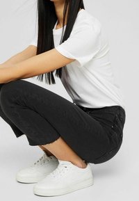 Camiseta blanca de manga corta, jeans negros hasta el tobillo con dobladillo deshilachado y zapatillas blancas. Diseño sencillo con una silueta ajustada.