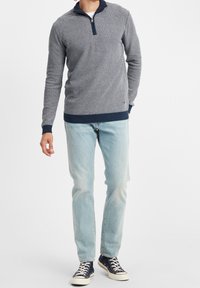 Pull en maille à motifs bleu foncé avec un col zippé, associé à un jean bleu clair et des baskets marine. Le pull a des poignets et un ourlet côtelés.