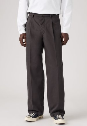 Homme portant un pantalon ample foncé, une chemise blanche à manches longues et des baskets noir et blanc, debout devant un fond clair.