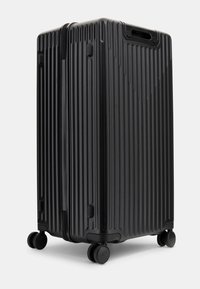 Maleta negra con ruedas, con un exterior estriado, diseñada para ser duradera. Presenta ruedas dobles giratorias y un mango retráctil para un fácil transporte.