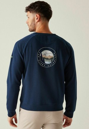 Mężczyzna w granatowej bluzie z grafiką góry i drzew oraz napisem „For Every Adventure” na plecach, stojący na jasnym tle.