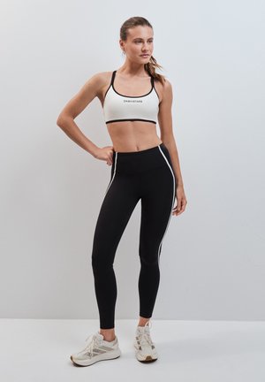 Donna in piedi con una mano sui fianchi, indossa un reggiseno sportivo bianco e nero, leggings neri con strisce bianche e scarpe da corsa bianche su sfondo semplice.