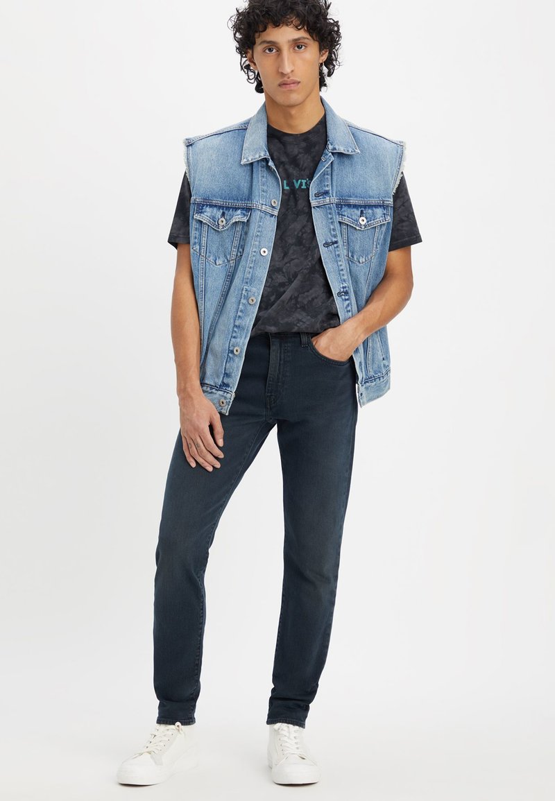 Gilet en denim de couleur bleue délavée, avec des découpes sur les épaules et deux poches avant, associé à un t-shirt noir à motif et un jean foncé. Baskets blanches.