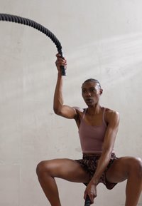 Atleet in een lichtroze tanktop en gestreepte shorts, die battle rope-oefeningen uitvoert in een neutraal gekleurde binnenruimte.
