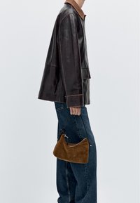 Brun läderjacka med en mockakrage, i kombination med vida blå jeans. En liten brun mockaväska hålls i vänster hand.