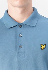 Blå bomullspolo skjorte med tre knapper i plassen, med et gult brodert ørne-logo på venstre bryst. Tekstureret stoff.