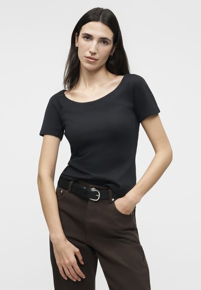 Γυναίκα με μακριά σκουρόχρωμα μαλλιά, που φοράει μαύρο τοπ με κοντά μανίκια, σκούρο καφέ παντελόνι και μαύρη ζώνη, στέκεται με το ένα χέρι στην τσέπη.