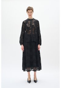 Schwarzes Spitzentop mit langen Ärmeln und floralem Muster, kombiniert mit einem passenden Rock. Mit Rundkragen und Knopfverschluss. Hochhackige Schuhe.