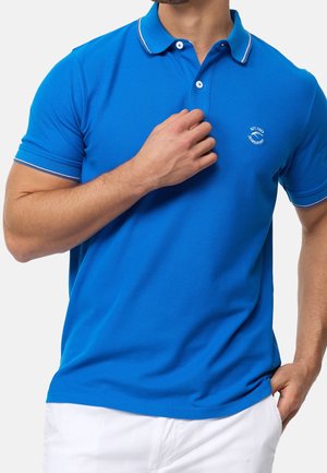 Man wearing a blue short-sleeve polo shirt with white trim on sleeves and collar, holding shirt near collar, white pants.