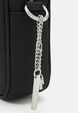 Coin de sac noir texturé avec chaîne argentée et étiquette rectangulaire Lacoste suspendue à une attache anneau argentée.