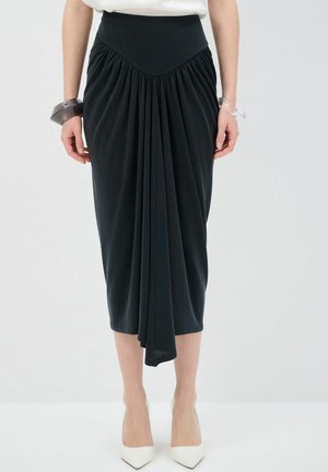 Jupe noire longueur genou en tissu lisse, dotée d'une taille haute et de détails froncés, associée à des escarpins blancs à bout pointu.