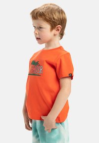 Oranžové bavlněné tričko s krátkými rukávy, s grafikou palm a textem "Quiksilver" v modré barvě. Nosené s tyrkysovými tie-dye šortkami.