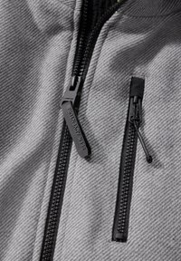 Veste grise texturée comportant deux poches zippées noires. Les tirettes de zip sont étiquetées "SEEK ADVENTURE". Le matériau semble durable et résistant aux intempéries.