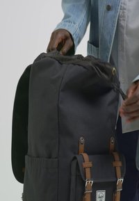 Svart tyg ryggsäck med bruna läderdetaljer, dragsko stängning, sidofickor och en främre klaff med spännband.