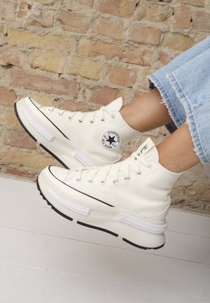 Białe wysokie trampki Converse noszone z krótkimi niebieskimi jeansami, pokazane na tle ceglastej ściany i białej podłogi.
