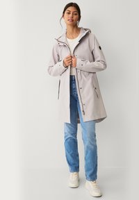Chaqueta impermeable gris claro con capucha, cremallera frontal y botones a presión; cuenta con dos bolsillos laterales y puños ajustables. Combinada con jeans azules.