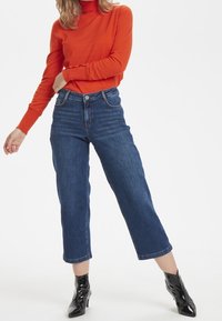 Jersey de cuello alto rojo y vaqueros oscuros de  cropped con corte recto. Botines de charol negros con puntera afilada. Diseño simple y limpio.