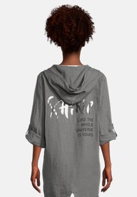 Veste à capuche en lin gris avec manches retroussées. Présente un texte imprimé blanc au dos, disant "BRILLE COMME SI L'UNIVERS ENTIER ETAIT À TOI."