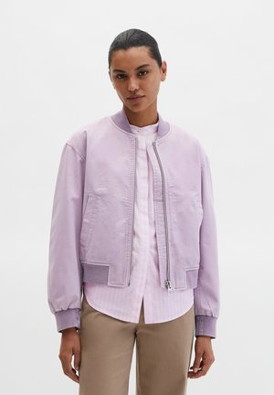 Lila Bomberjacke mit gerippten Bündchen und Saum, ausgestattet mit einem Frontreißverschluss und Seitentaschen. Trägt man über einem hellrosa gestreiften Hemd.
