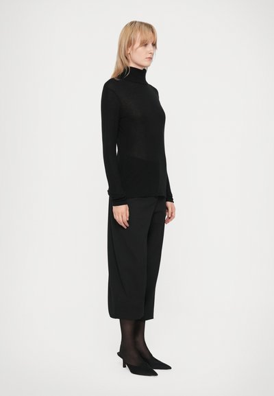 Crna turtleneck majica od laganog materijala, uparena s širokim crnim culottes hlačama i špicastim crnim potpeticama, nosi stajajući lik.