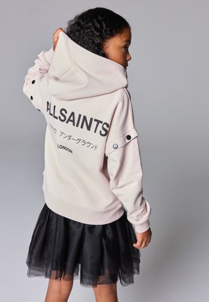 Junges Mädchen trägt einen blassen Hoodie mit dem Schriftzug "ALLSAINTS" und einen schwarzen Rock, hält die Kapuze mit einer Hand und blickt nach rechts auf grauem Hintergrund.