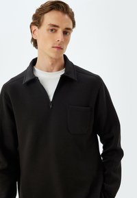Pullover zip-up nero realizzato in un tessuto morbido, con una tasca frontale, colletto e maniche lunghe. Il design è semplice e moderno.