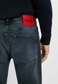Jeans in denim scuro con una finitura scolorita, caratterizzati da una toppa rettangolare rossa con la scritta "HUGO" in nero e tasche posteriori standard.