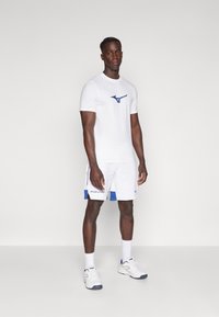 Bala sporta t-krekli ar zilu logotipu, apvienoti ar baltām šortiem, kas izceļas ar ziliem akcentiem un tekstu. Apavi ir baltā krāsā ar ziliem akcentiem.