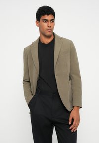 Khaki blazer met notch lapels, textuurstof, enkele knoopsluiting en zijzakken, gecombineerd met een zwart T-shirt en donkere broeken.