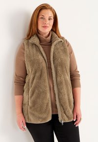 Fuzzy beige väst med hög krage, dragkedjestängning och sidofickor, lager på en brun polotröja, kombinerad med svarta byxor.