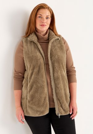 Fuzzy beige vest met hoge kraag, ritssluiting en zijzakken, gelaagd over een bruine coltrui, gecombineerd met zwarte broek.