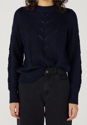 Femme portant un pull en maille torsadée bleu marine rentré dans un jean noir taille haute sur un fond uni.