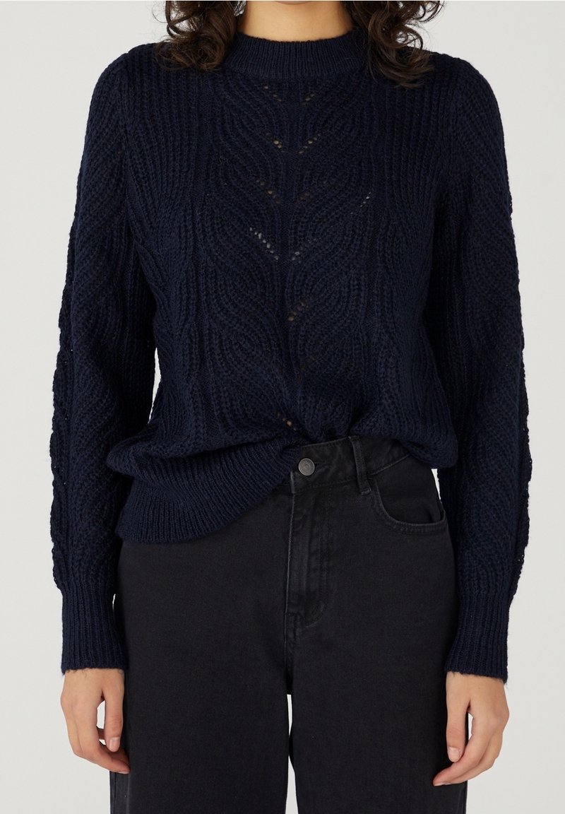 Femme portant un pull en maille torsadée bleu marine rentré dans un jean noir taille haute sur un fond uni.
