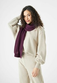 Jonge vrouw met krullend haar, gekleed in beige loungewear en een paarse gebreide sjaal, poseert met één hand achter haar hoofd.