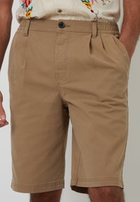 Beige Shorts aus einem Baumwoll-Blend-Gewebe, mit Knopfverschluss, verstellbarem Bund und Seitentaschen. Glatte Textur mit gefälteltem Design.
