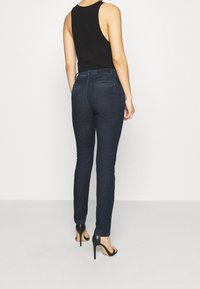 Pantalones vaqueros skinny de denim oscuro con una cintura de altura media, que presentan dos bolsillos traseros y una textura suave, combinados con sandalias negras de tacón alto.