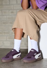 Lila mockaskor med nättyg och vit branding. De har en texturerad gummisula och är tillsammans med vita strumpor och beige ribbade byxor.