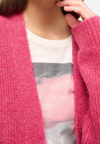 Rosa stickad cardigan med ribbade manschetter, lager på lager över en ljus grafisk t-shirt med abstrakt rosa och grå design. Mjuk textur, avslappnad passform.