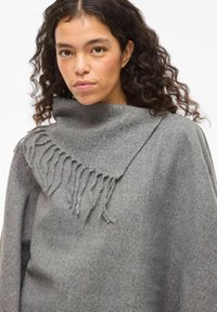 Sieviete ar cirtainiem matiem, kura valkā pelēku vilnas šalli un atbilstošu šaltu ar frēzēšanas detaļām.