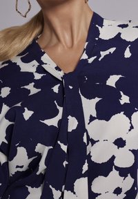 Blouse marine avec un motif floral abstrait blanc, dotée d'un décolleté en V et de plis à l'avant. Texture de tissu lisse.
