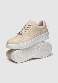 Béžové tenisky s embosovaným vzorom loga, šnurovací dizajn, polstrovaný golier a textúrovaná biela gumová podrážka s obvodovými detailmi.