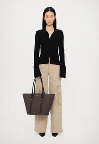 Brun mønstret tote bag med sorte læderhåndtag, båret med en sort ribbet skjorte med knapper og beige cargo bukser, der har sidelommer.