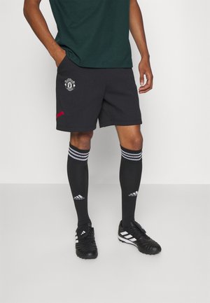 Homme portant un short noir de Manchester United, des chaussettes noires Adidas montantes jusqu'aux genoux et des chaussures de football Adidas noires, debout sur un fond blanc.