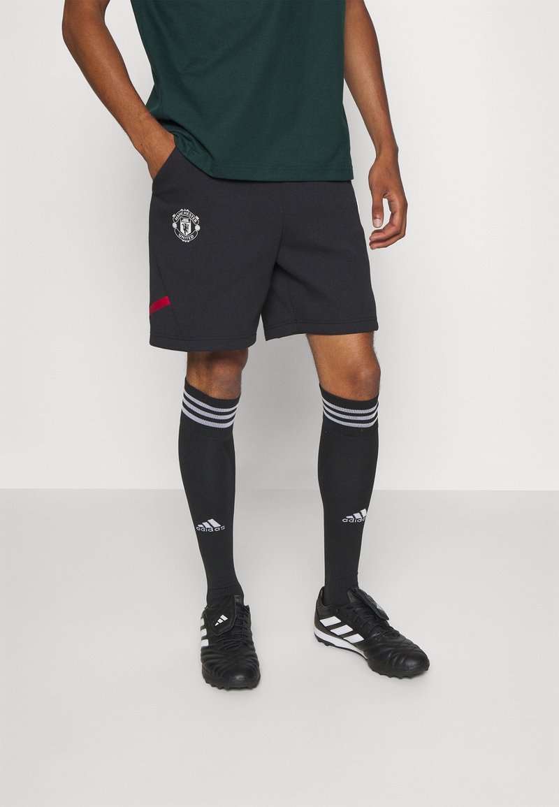 Homme portant un short noir de Manchester United, des chaussettes noires Adidas montantes jusqu'aux genoux et des chaussures de football Adidas noires, debout sur un fond blanc.