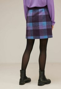 En person står med ryggen mot kameran, iklädd en lila topp, en rutigt kjol i blå och lila nyanser, samt svarta ankelstövlar.