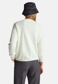 Cremfarbener Strickpullover mit gerippten Bündchen und Saum, kombiniert mit taillierten Hosen in Anthrazit. Ein schwarzer Bucket Hat setzt einen kontrastierenden Akzent.