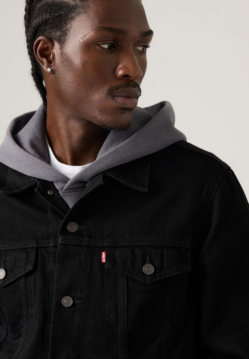 Black denim jacket with two chest pockets, silver buttons, and a red logo tab; layered over a gray hooded sweatshirt.