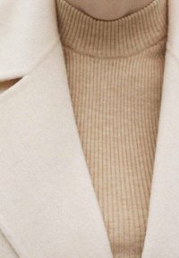 Suéter de cuello alto de canalé beige, superpuesto bajo un abrigo de lana crema con solapa ancha, que presenta una textura suave y un aspecto limpio y ajustado.