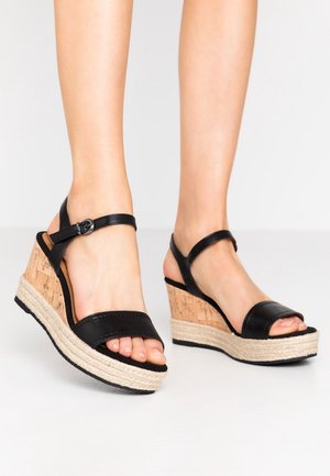 Sorte kilehøje med kork-mellemsål og jute-detaljer. Har rem om anklen og åben tå, der fremhæver de teksturerede materialer.