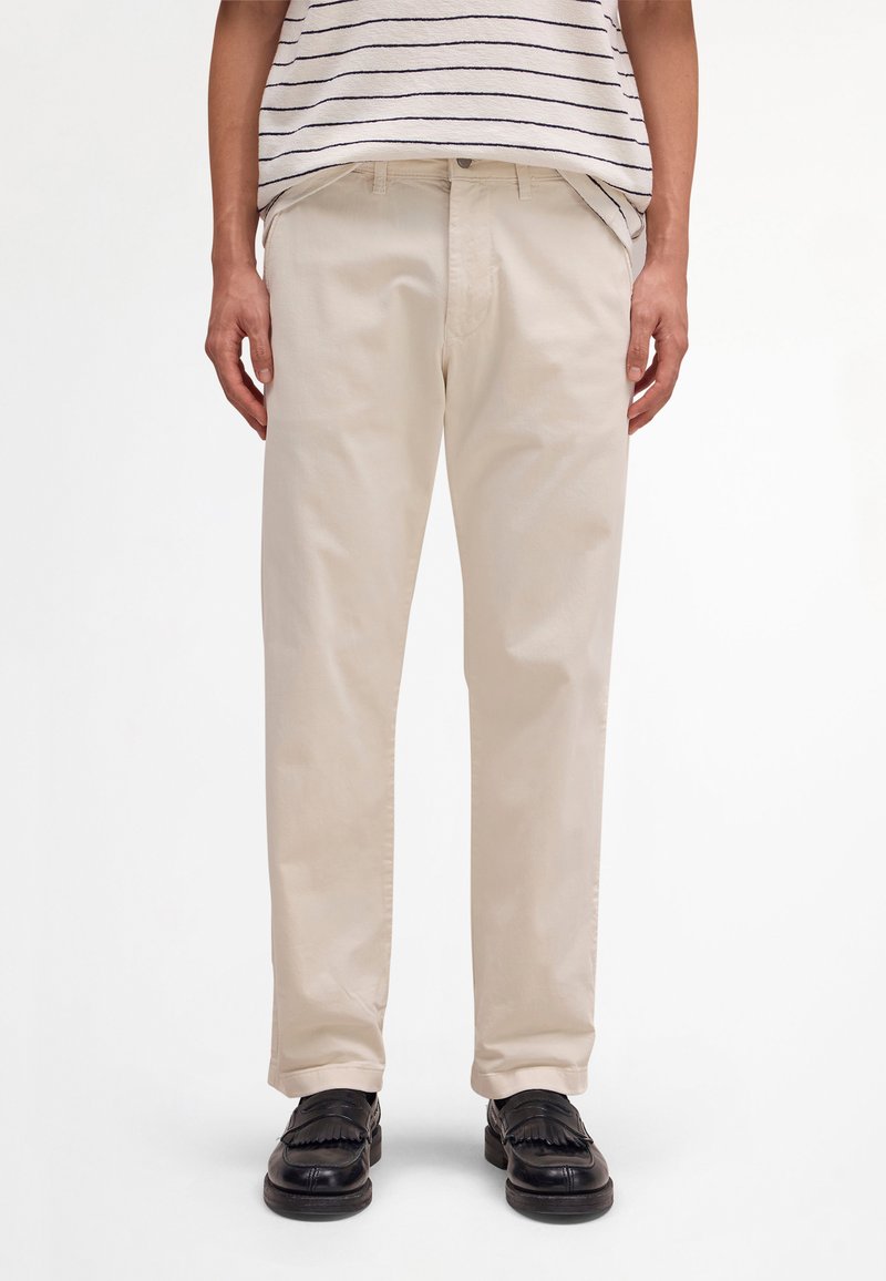 Mann, der beige gerade geschnittene Hosen, schwarze Slipper und ein weißes Hemd mit dünnen schwarzen horizontalen Streifen trägt.